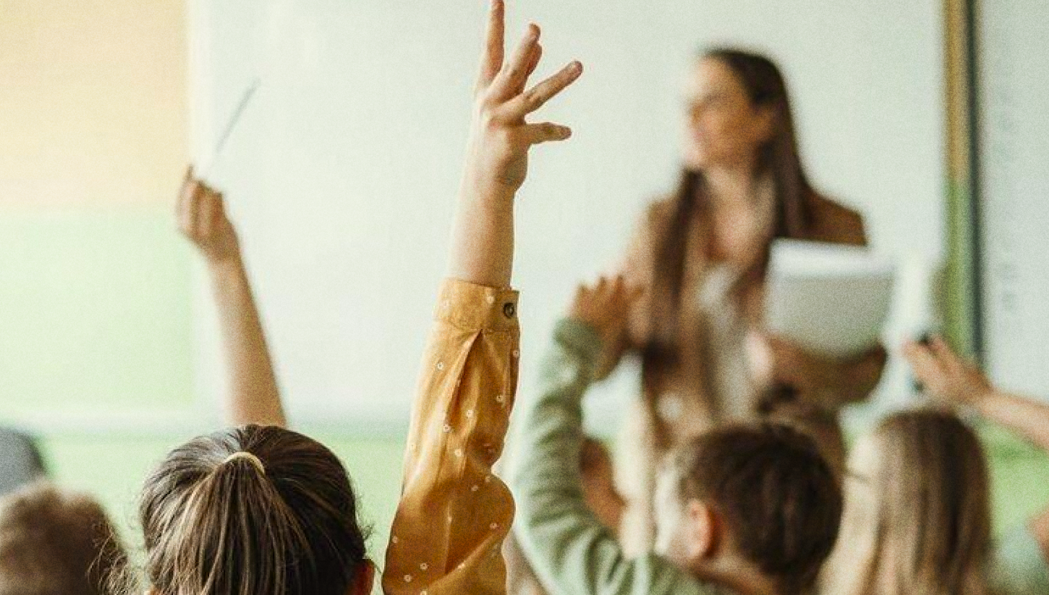Crianças sentadas em sala de aula levantam as mãos diante de uma professora.