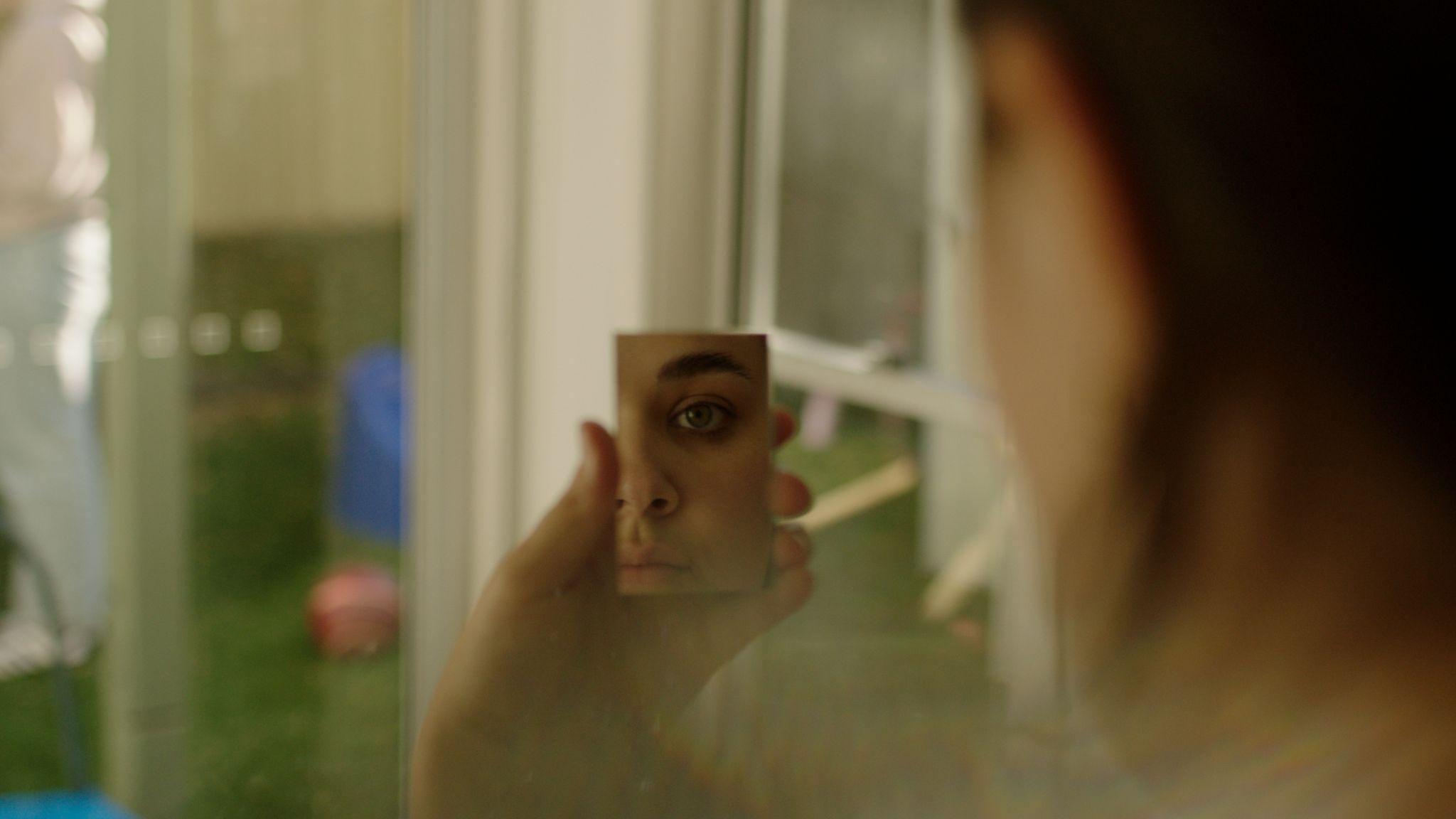 Pessoa observa o próprio reflexo em um pequeno espelho em cena do documentário Olhar pra Si